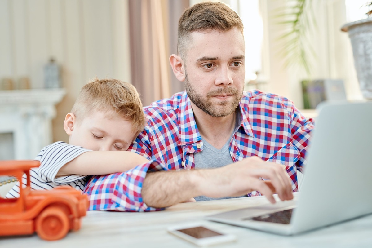 Warm toned portrait of cute little boy falling asleep hugging his dad waiting for him to finish working at laptop; remote work challenges concept