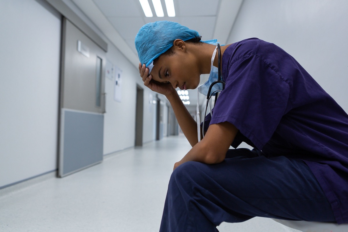 Side view of tense female surgeon with hand on forehead; covid unions and mental health concept