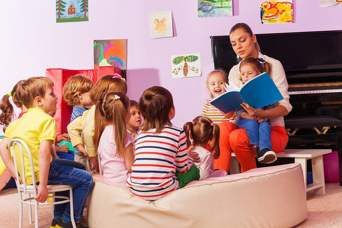 Large group of kids sit and listen to teacher reading a book and telling stories; teacher shortage concept