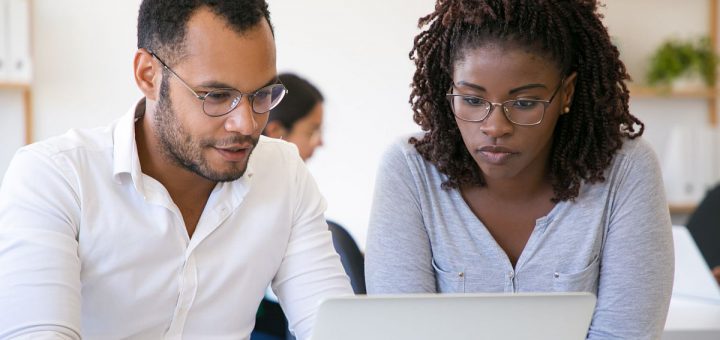 Diverse colleagues watching content on laptop together. Young man and woman using computer in office, looking at screen and talking. Corporate discussion concept