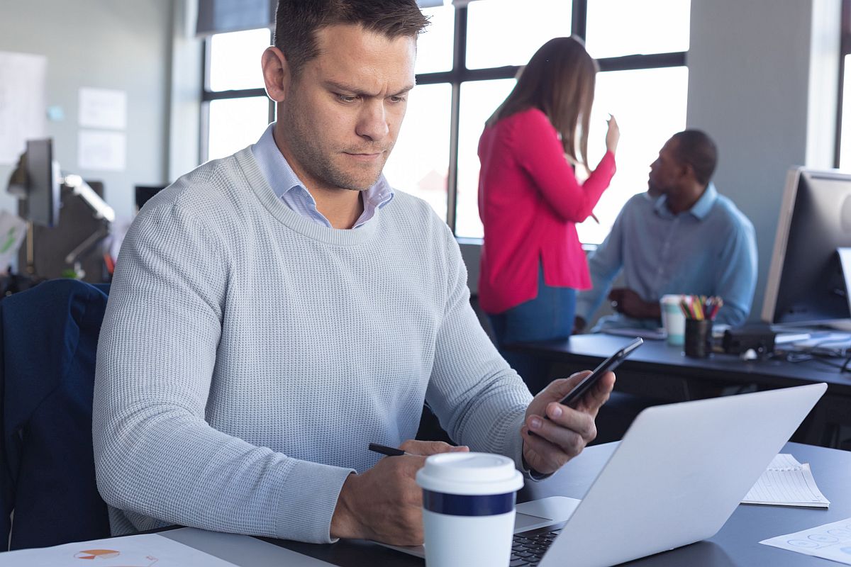 Man working in a casual modern office, using a laptop and looking at his smartphone, with colleagues in the background; union-busting during the union drive concept