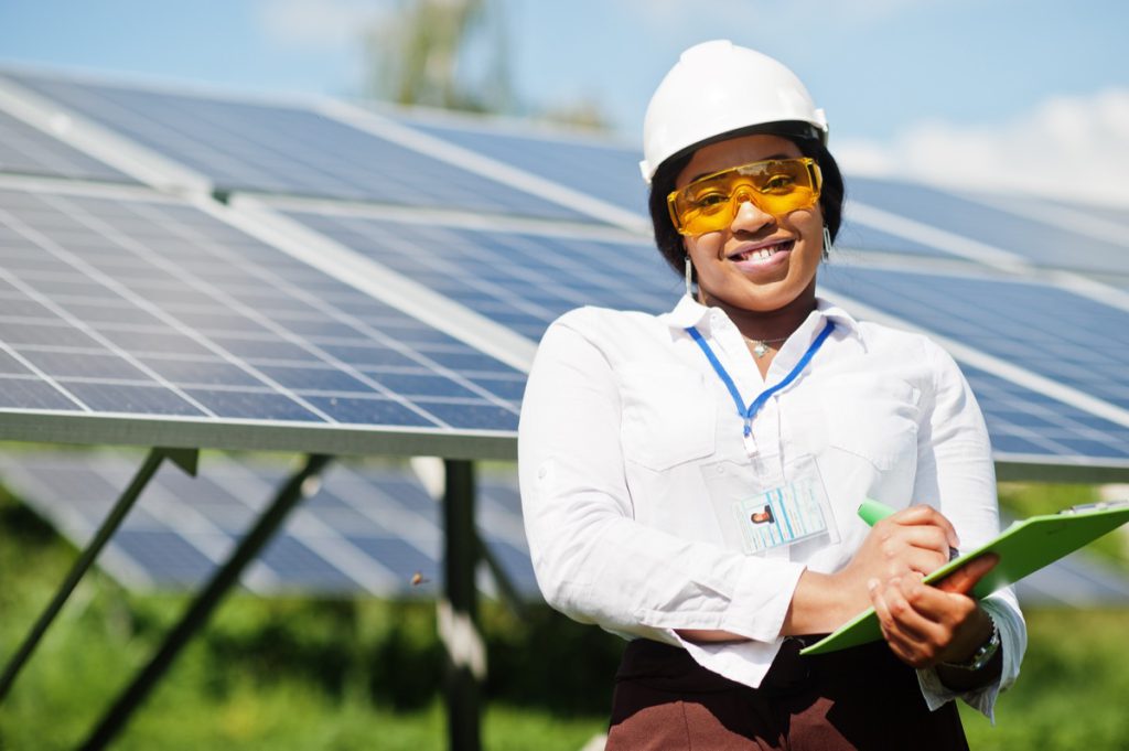 Woman wearing white hard hat, holding a tablet, stands in front of solar panels; Inflation Reduction Act benefits concept